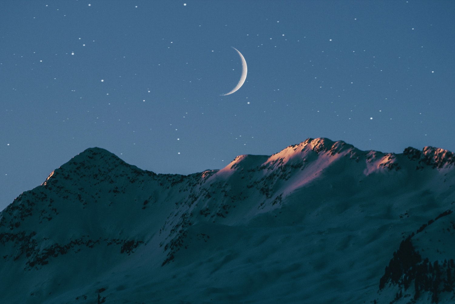 Very thin crescent Moon in a night sky, just rising above rocky mountains